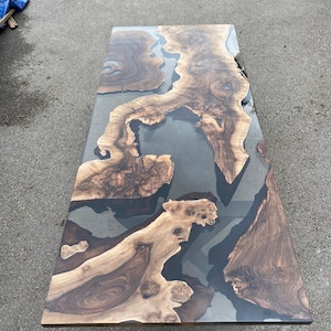 Puede incluir: Una mesa rectangular de madera con un acabado de resina epoxi transparente. La mesa presenta patrones de vetas de madera natural en tonos marrones y beige, con resina gris oscuro que rellena los espacios entre la madera. La mesa mide aproximadamente 183 cm de largo.