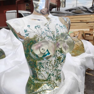 Puede incluir: Una escultura de busto de vidrio transparente, con arreglos florales internos. La escultura presenta orqu&iacute;deas blancas y moradas claras, junto con un delicado follaje verde y azul. El busto se exhibe sobre una superficie blanca, con un fondo borroso de un edificio y palets de madera.