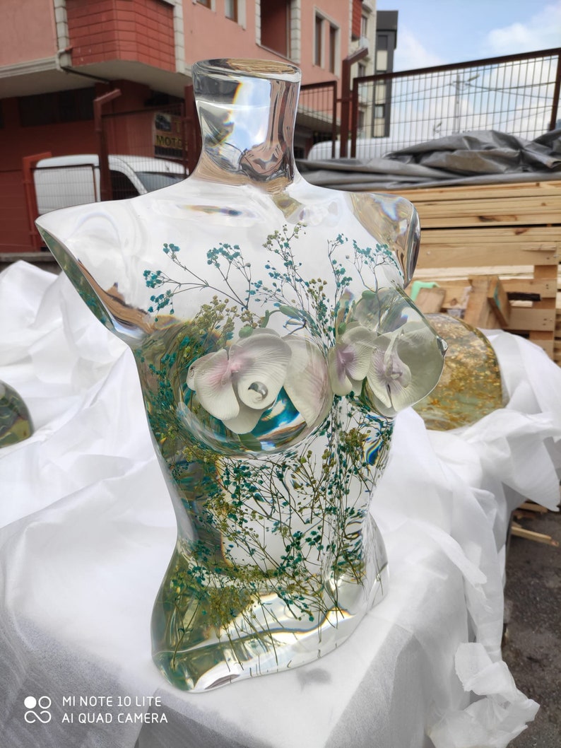 Puede incluir: Una escultura de busto de vidrio transparente, con arreglos florales internos. La escultura presenta orqu&iacute;deas blancas y moradas claras, junto con un delicado follaje verde y azul. El busto se exhibe sobre una superficie blanca, con un fondo borroso de un edificio y palets de madera.