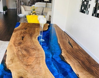 May include: A handcrafted wooden table with a vibrant blue epoxy river running through the center. The table features natural wood grain and a glossy finish. The table is approximately 96 inches long.