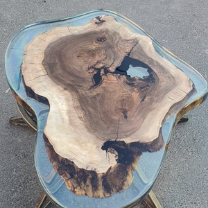 May include: A unique coffee table with a live edge wood slab encased in clear resin. The wood features natural grain patterns and dark knots. The table has a clear resin top and brass-colored legs, creating a modern, organic aesthetic.