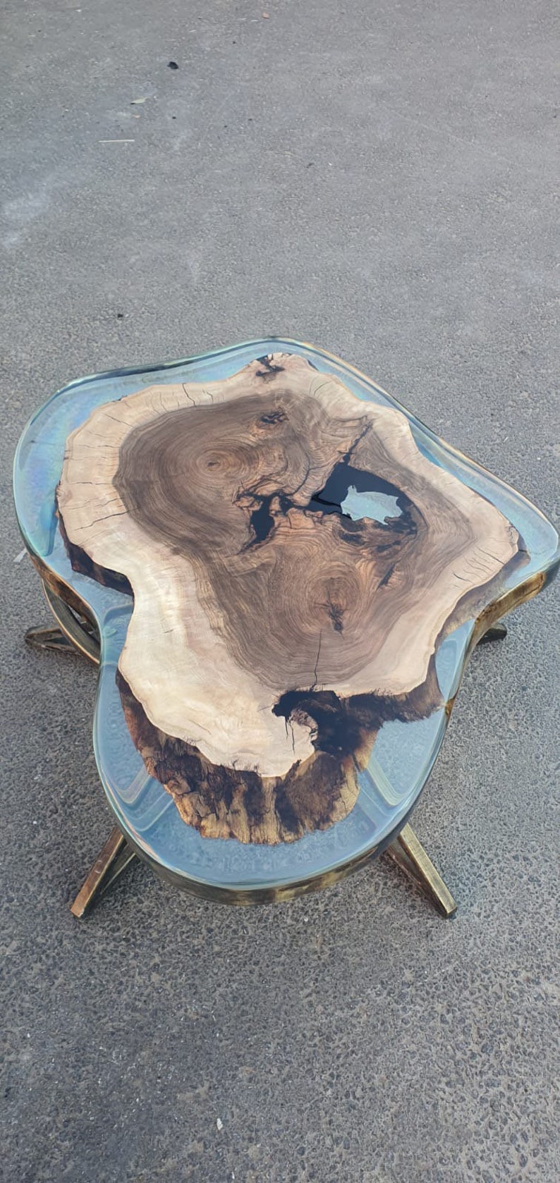 May include: A unique coffee table with a live edge wood slab encased in clear resin. The wood features natural grain patterns and dark knots. The table has a clear resin top and brass-colored legs, creating a modern, organic aesthetic.