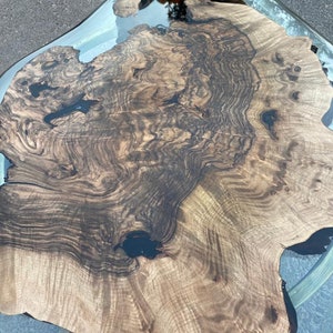 May include: A unique, handcrafted coffee table with a live edge wood slab top. The wood features a rich, dark brown grain pattern and is encased in a clear resin. The table has a modern design and is supported by a metal frame.