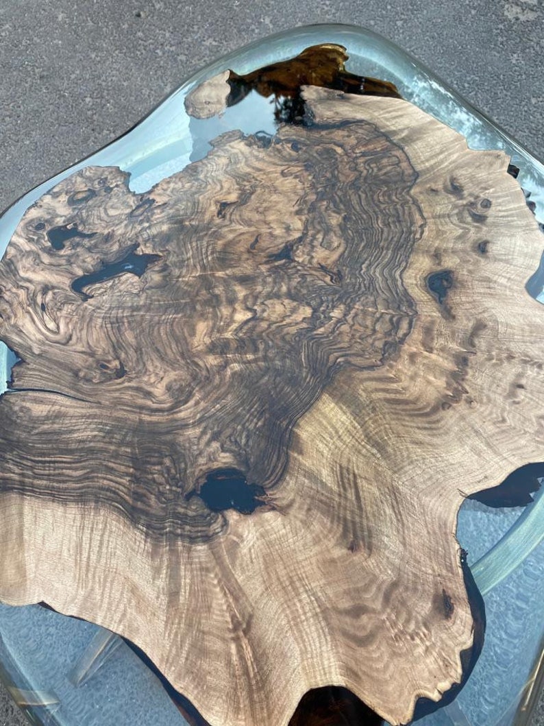 May include: A unique, handcrafted coffee table with a live edge wood slab top. The wood features a rich, dark brown grain pattern and is encased in a clear resin. The table has a modern design and is supported by a metal frame.