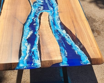 Puede incluir: Mesa de madera artesanal con un río de resina epoxi azul en el centro. La mesa tiene un acabado de vetas de madera natural y una base de metal negro. El diseño del río de resina epoxi presenta detalles blancos.