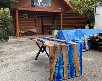 May include: A handcrafted wooden table with a blue resin river design. The table features a natural wood grain and is supported by black metal legs. The table is outdoors in front of a wooden building with the text "Lara Wood's".