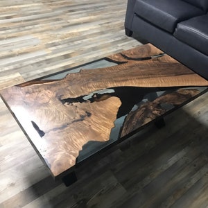 May include: A rectangular coffee table with a unique design. The table features a wooden surface with a natural grain pattern, filled with a dark epoxy resin. The table is supported by black metal legs. The table is in a living room setting.