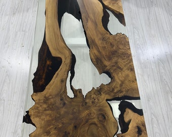 Puede incluir: Una mesa de centro rectangular con una tapa de cristal transparente y una base de madera única. La base presenta un patrón de vetas de madera natural con tonos marrón oscuro y marrón claro, y resina negra rellena los huecos. Las dimensiones de la mesa no son visibles.