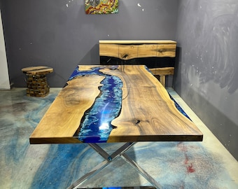 May include: A large wooden dining table with a blue resin river design. The table has a natural wood grain finish and sits on a polished metal base. A matching wooden cabinet is in the background.