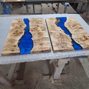 May include: Two wooden slabs with a river of blue resin running through them. The wood has a light brown, natural grain. The slabs are on a white surface in a workshop setting.