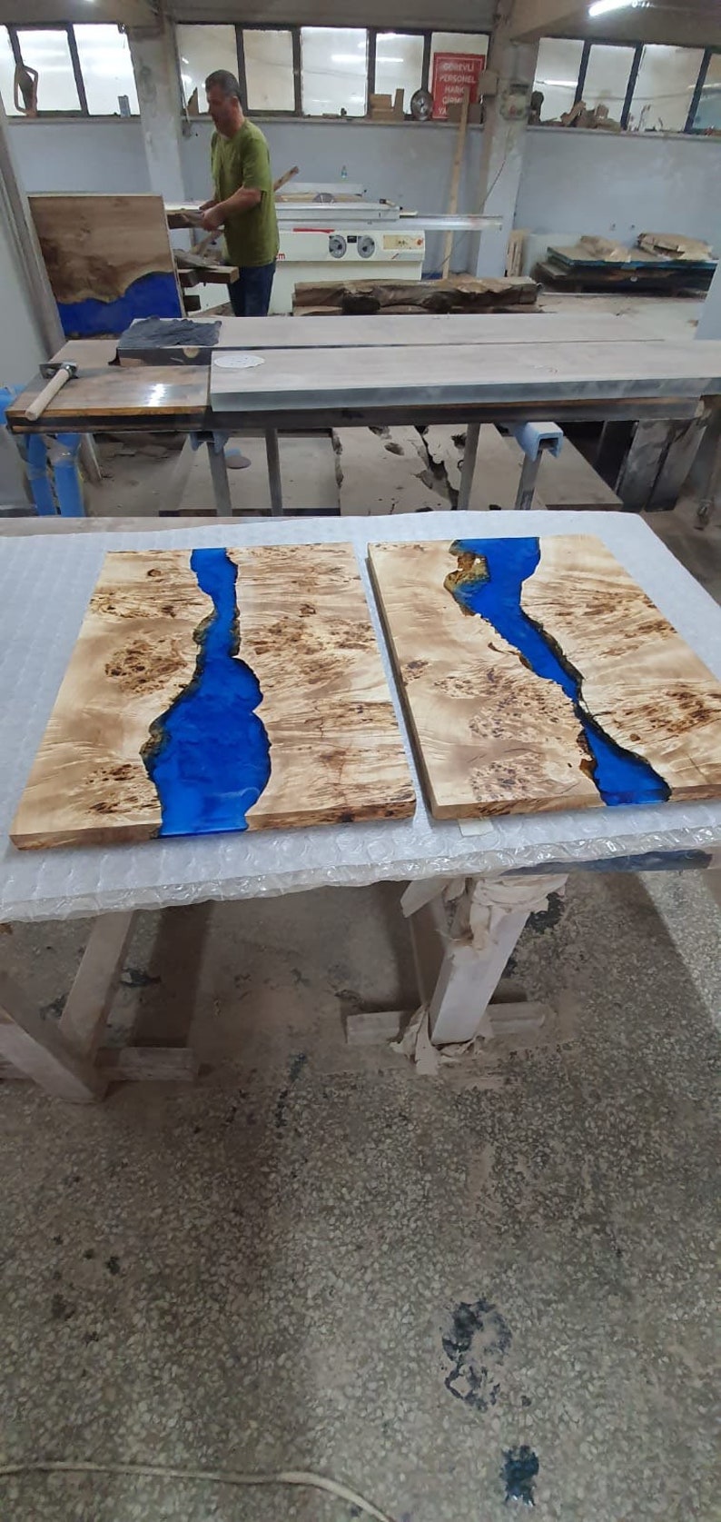 May include: Two wooden slabs with a river of blue resin running through them. The wood has a light brown, natural grain. The slabs are on a white surface in a workshop setting.
