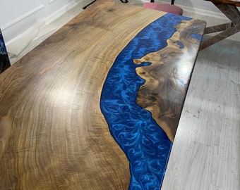 May include: A large, rectangular wooden table with a river of blue resin flowing through the center. The wood has a natural grain pattern. Other tables are visible in the background, showcasing a variety of designs and styles.