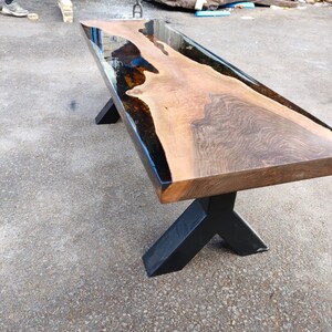 May include: A long, rectangular wooden table with a dark, glossy epoxy resin river running through the center. The table has a natural wood grain finish and sits on black metal legs. The table is approximately 183 cm long.