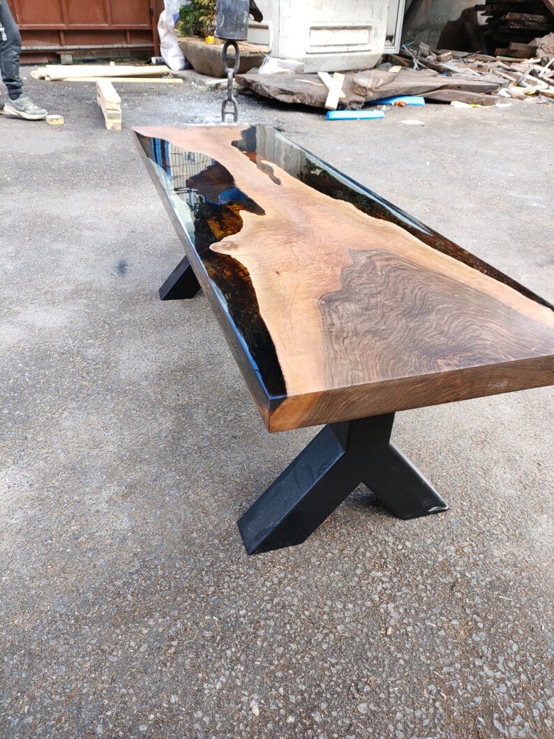 May include: A long, rectangular wooden table with a dark, glossy epoxy resin river running through the center. The table has a natural wood grain finish and sits on black metal legs. The table is approximately 183 cm long.