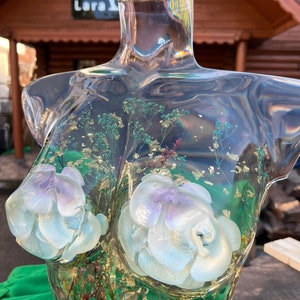 Puede incluir: Escultura de busto de resina transparente con elementos florales blancos y lavanda incrustados y detalles de pan de oro. El busto presenta dos grandes formas de flores esculpidas y varias flores secas. La escultura se exhibe al aire libre.