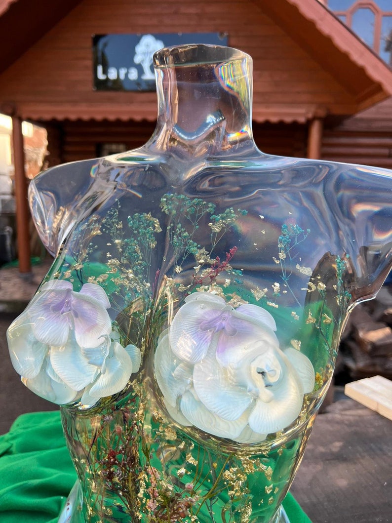 Puede incluir: Escultura de busto de resina transparente con elementos florales blancos y lavanda incrustados y detalles de pan de oro. El busto presenta dos grandes formas de flores esculpidas y varias flores secas. La escultura se exhibe al aire libre.
