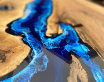 May include: A close-up of a wooden table with a river of blue resin. The wood has a natural grain and a light brown color. The blue resin flows through the center, creating a striking contrast. The resin has a translucent, glowing effect.