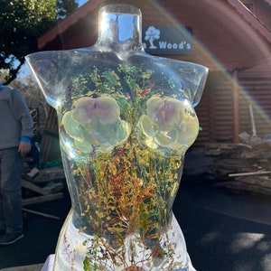 Puede incluir: Una escultura de resina transparente con forma de torso, llena de flores secas, plantas y una concha. La escultura tiene dos grandes formas de flores claras incrustadas en el &aacute;rea del pecho. La escultura se exhibe al aire libre.