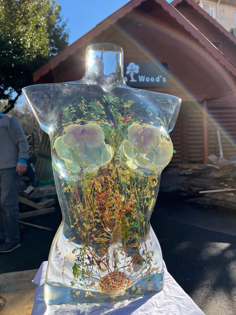Puede incluir: Una escultura de resina transparente con forma de torso, llena de flores secas, plantas y una concha. La escultura tiene dos grandes formas de flores claras incrustadas en el &aacute;rea del pecho. La escultura se exhibe al aire libre.