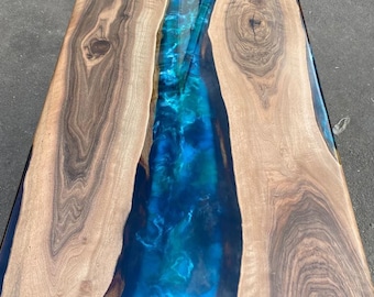 Puede incluir: Mesa rectangular de madera con un río de resina epoxi azul en el centro. La madera tiene un veteado natural y es de color marrón claro. La resina es una mezcla de azules y verdes.