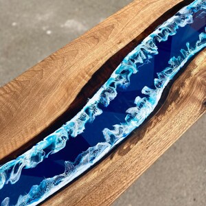 May include: A wooden table with a river of blue resin running through the center. The resin has white and light blue wave-like patterns along the edges, creating a coastal theme. The wood has a natural grain.