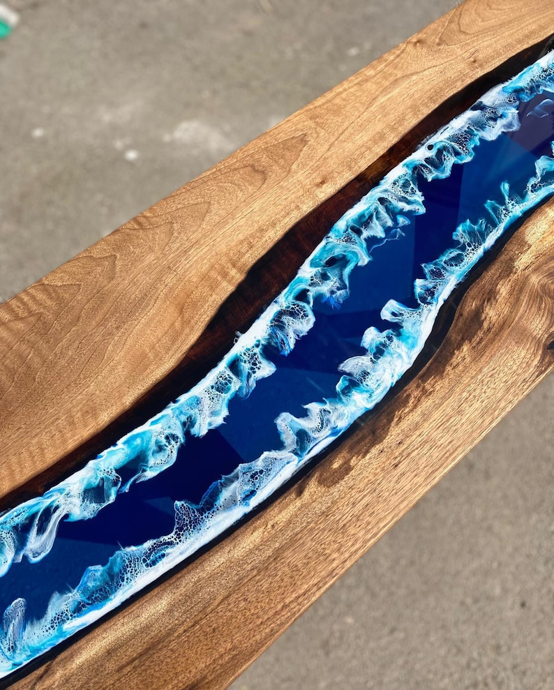 May include: A wooden table with a river of blue resin running through the center. The resin has white and light blue wave-like patterns along the edges, creating a coastal theme. The wood has a natural grain.