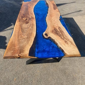 May include: A handcrafted wooden table with a striking blue epoxy resin river. The table features a natural wood grain finish and is supported by black metal legs. The table's dimensions are approximately 72 inches long and 36 inches wide.