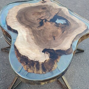 May include: A unique coffee table with a live edge wood top and a clear epoxy resin border. The wood has a natural grain pattern with dark and light brown tones. The table is supported by brass-colored metal legs.