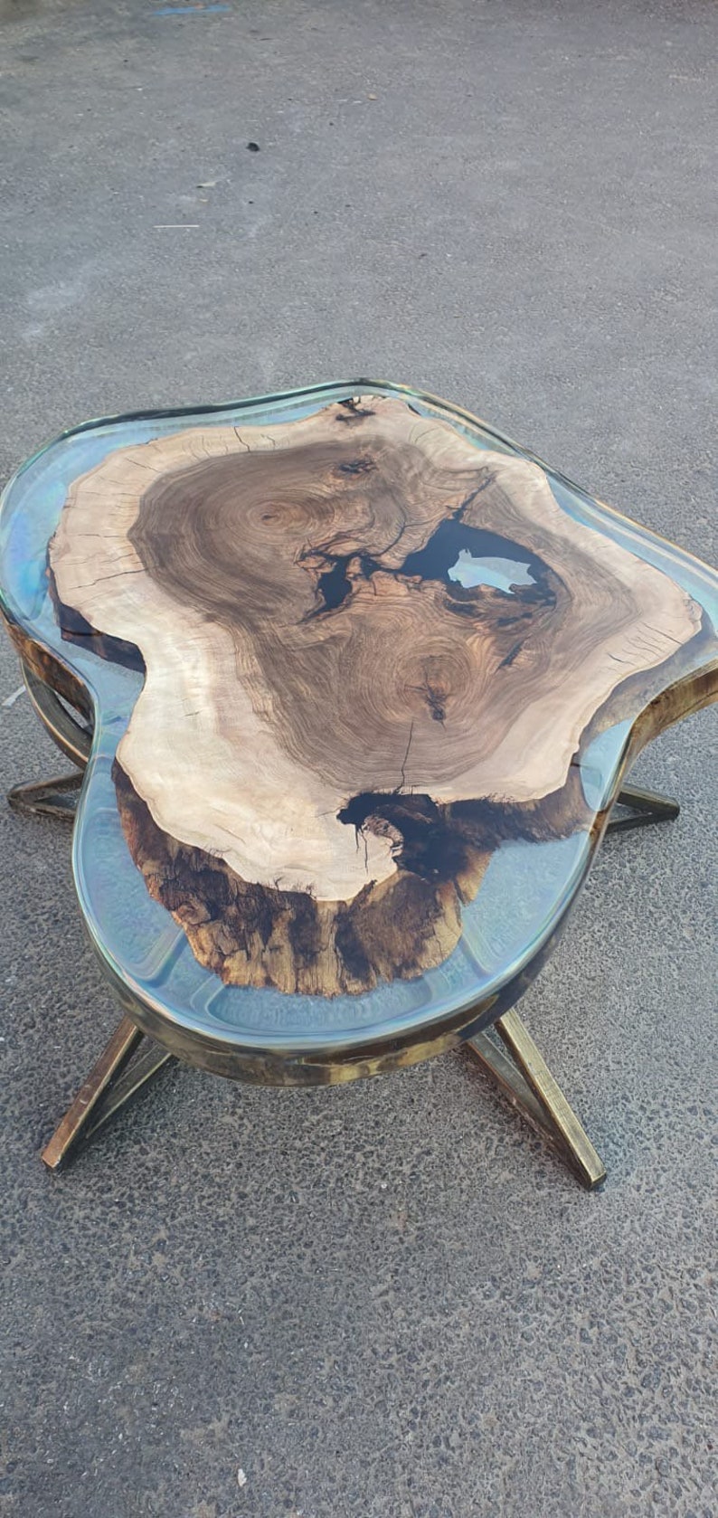 May include: A unique coffee table with a live edge wood top and a clear epoxy resin border. The wood has a natural grain pattern with dark and light brown tones. The table is supported by brass-colored metal legs.