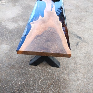 May include: A rectangular wooden table with a live edge and a dark blue epoxy resin fill. The table has a black, X-shaped base. The wood grain is visible. The table is approximately 152 cm long.