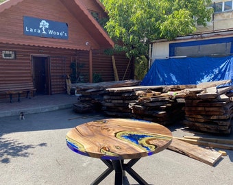 May include: A round wooden table with a unique epoxy resin design. The tabletop features natural wood grain with blue and gold resin accents. The table has a black metal base and is displayed outdoors near a building with the sign "Lara Wood's".