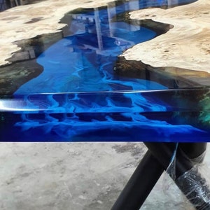 Op de afbeelding: Een handgemaakte houten tafel met een blauw epoxyhars rivierontwerp. De tafel heeft een natuurlijke houtnerf aan de randen en een glanzend, reflecterend oppervlak. De blauwe hars creëert een vloeiend, rivierachtig effect. Zwarte metalen poten.