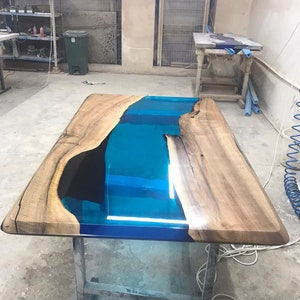 May include: A rectangular wooden table with a river of blue resin running through the center. The table has a natural wood grain finish on the edges and a glossy, reflective surface. The table is supported by a metal frame.