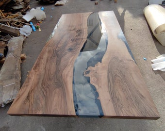 Könnte beinhalten: Großer, rechteckiger Holztisch mit einem dunklen, glänzenden Epoxidharzfluss in der Mitte. Das Holz hat ein natürliches Maserungsmuster mit Braun- und Beigetönen. Das Harz reflektiert den Himmel. Der Tisch ist unfertig.