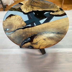 May include: A round wooden table with a unique design. The tabletop features a combination of light brown wood and a clear, blue-tinted resin. The table is supported by a dark metal base. The table measures approximately 40 inches in diameter.