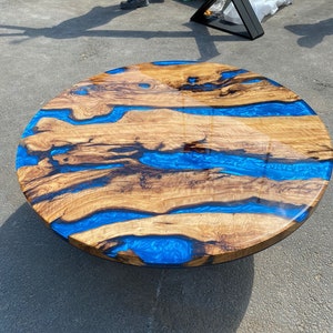 May include: Round wooden table with a unique design. The tabletop features natural wood grain with vibrant blue resin accents, creating a river-like effect. The table is a light brown color with a glossy finish. The table is approximately 48 inches in diameter.