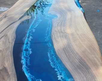 May include: A large, rectangular wooden table with a blue epoxy resin river design. The wood has a natural grain, and the resin mimics ocean waves. The table is likely for a dining or living room.