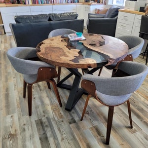 May include: A round wooden table with a live edge and a blue resin inlay, surrounded by four gray upholstered chairs with wooden legs. The table has a black metal base. The room has a bookshelf and a gray sofa.