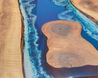 May include: A handcrafted wooden table with a river-like design. The table features a blue epoxy resin center, resembling water, and is surrounded by natural wood. The table is supported by a dark metal base.