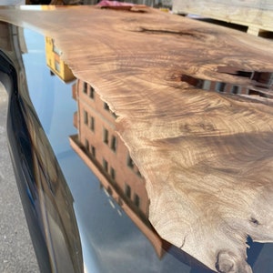 May include: A live edge wooden table with a dark, glossy finish. The wood grain is visible, showcasing natural patterns and textures. The table reflects the surrounding environment, including a building and the sky.
