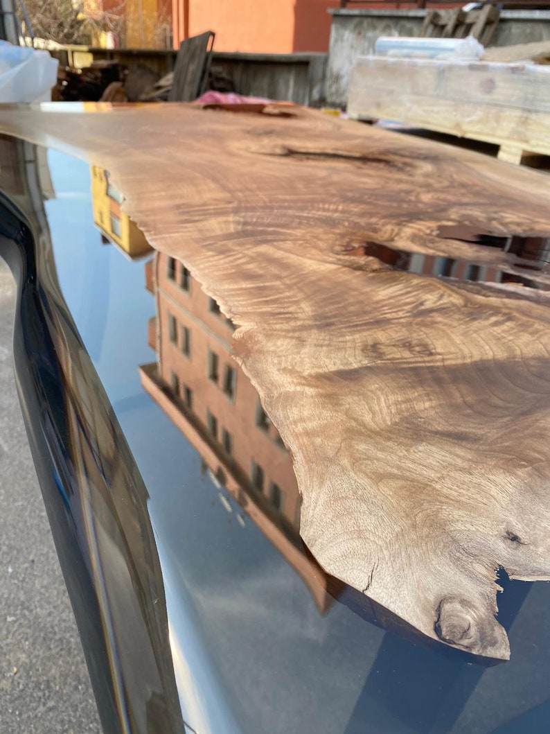 May include: A live edge wooden table with a dark, glossy finish. The wood grain is visible, showcasing natural patterns and textures. The table reflects the surrounding environment, including a building and the sky.