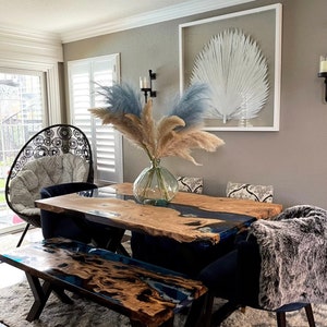 May include: A dining room scene featuring a live edge wooden table and bench with blue resin accents. A clear glass vase holds pampas grass. A framed white palm leaf art piece hangs on the gray wall. A black chair with a patterned cushion is visible.