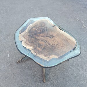 May include: A unique coffee table with a live edge wood top encased in a clear resin. The wood grain is visible, and the table is supported by a bronze-colored metal base. The table's dimensions are approximately 40 inches long and 24 inches wide.