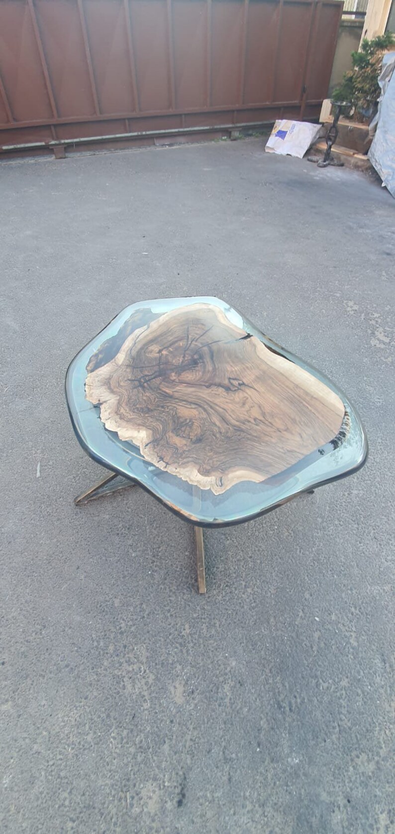 May include: A unique coffee table with a live edge wood top encased in a clear resin. The wood grain is visible, and the table is supported by a bronze-colored metal base. The table's dimensions are approximately 40 inches long and 24 inches wide.