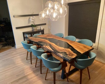 Op de afbeelding: Een grote, op maat gemaakte eettafel met een houten blad met levende rand en een heldere epoxyharsrivier. De tafel wordt omringd door acht turquoise stoelen met houten poten. Een moderne kroonluchter hangt boven de tafel.