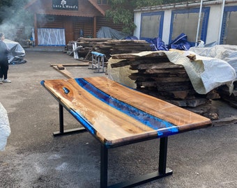 May include: A rectangular wooden table with a blue resin river design. The tabletop is supported by black metal legs. The table appears to be outdoors, with a wooden building in the background.