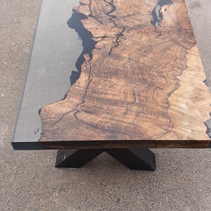 May include: Rectangular wooden coffee table with a dark epoxy resin inlay. The wood grain is visible, showcasing a natural pattern. The table has a black metal X-shaped base. The table's dimensions are approximately 60 inches long and 30 inches wide.
