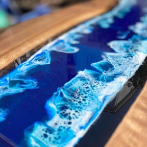 May include: A handcrafted wooden serving board with a deep blue epoxy resin design resembling ocean waves. The resin features white and light blue accents, creating a coastal aesthetic. The board's wooden edge is a warm brown color.