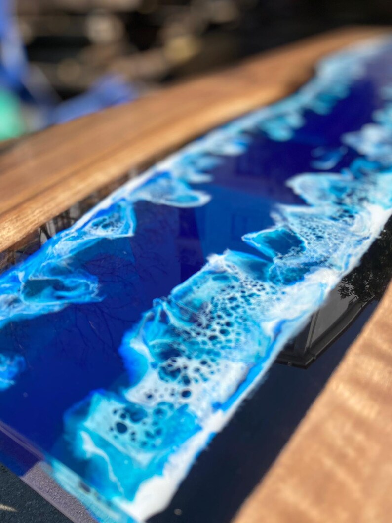 May include: A handcrafted wooden serving board with a deep blue epoxy resin design resembling ocean waves. The resin features white and light blue accents, creating a coastal aesthetic. The board's wooden edge is a warm brown color.