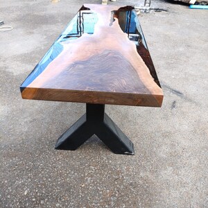 May include: A large, rectangular wooden table with a live edge and a glossy, reflective surface. The table is supported by a black, angular metal base. The wood grain is visible, showcasing natural patterns.